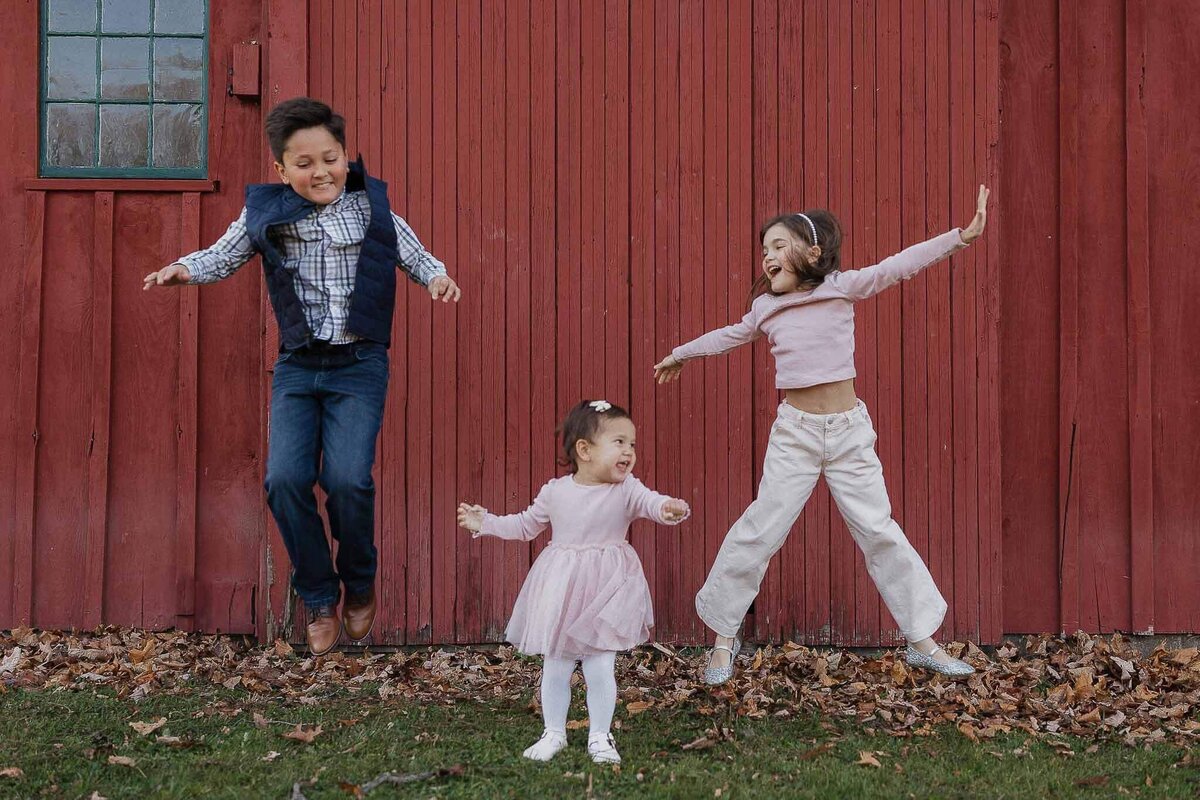 Three siblings jumping in front of a red bard during Christmas mini session at Sugar Pines Tree Farm in Chesterland, Ohio.