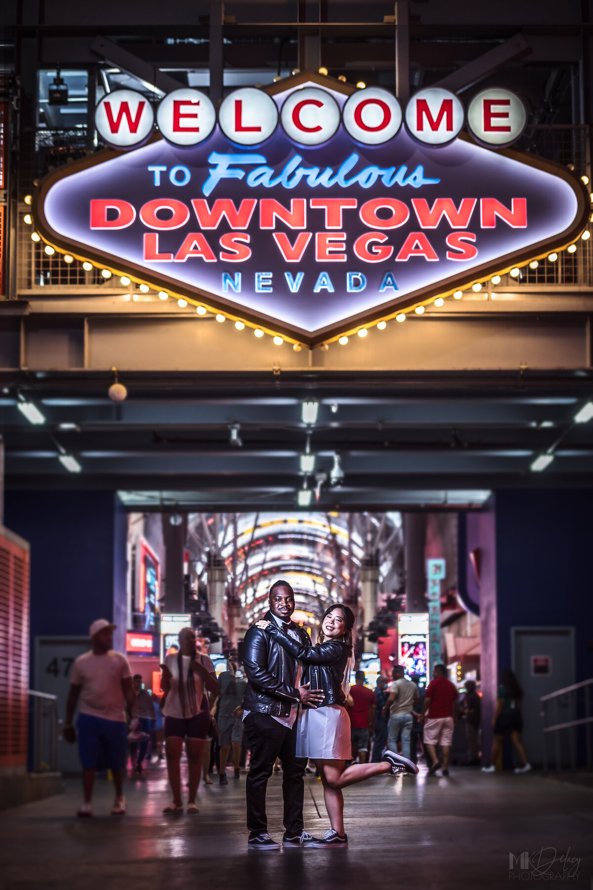 Welcome to Fabulous  Downtown  Las Vegas  sign on fremont bride holding groom with her foot popped while groom holds her at waist and they smile at camera under sign bride in white dress with black leather jacket grom in skinny jeans and white dress shirt with black leather jacket wearing  converse on Fremont st  las vegas elopement eloping in vegas  las vegas wedding photographers las vegas wedding photography mk delacy photography
