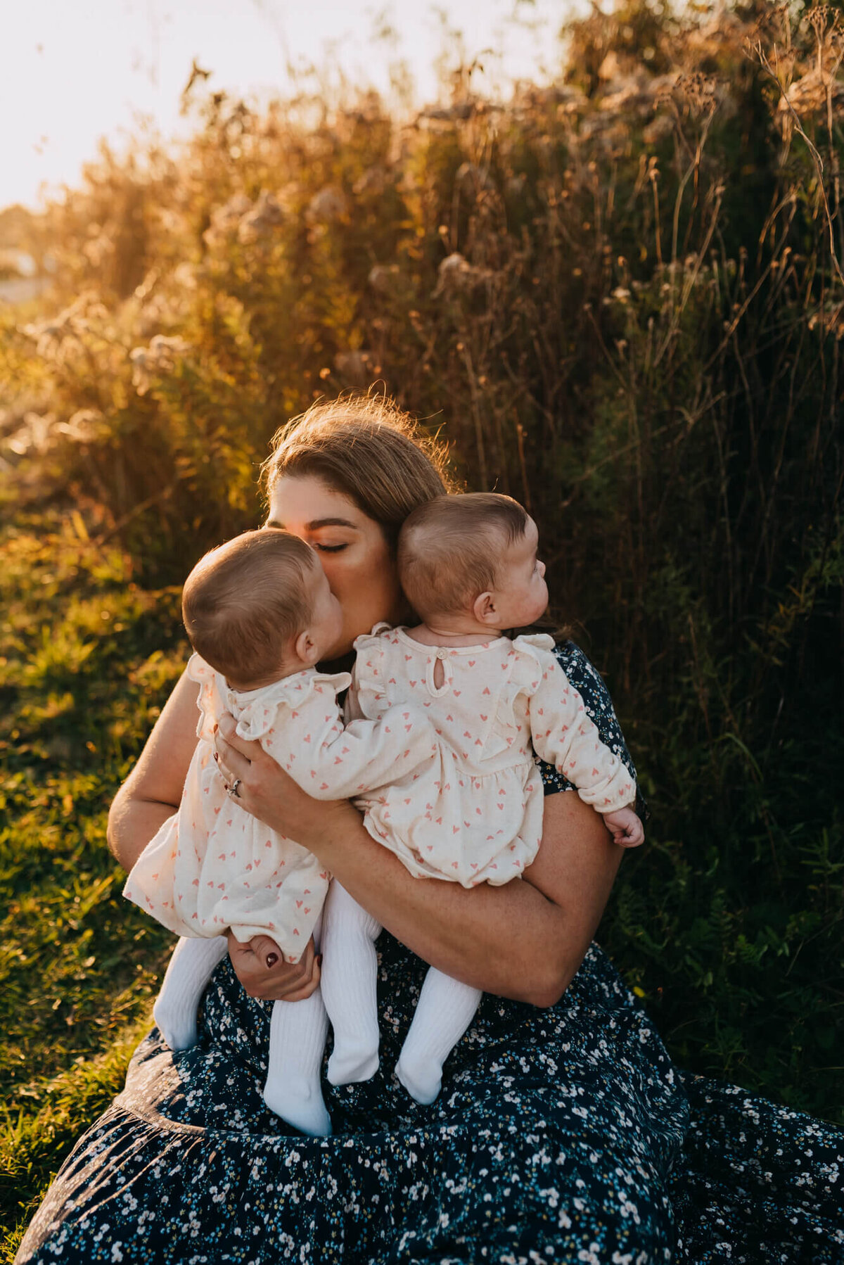 A mother in a blue and white floral dress sits on the ground and snuggles her twin daughters at sunset
