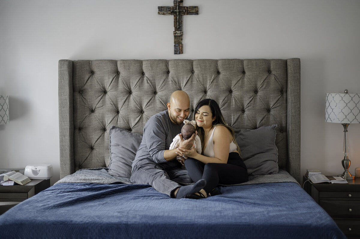 Parents sit in bed while holding their newborn daughter as they look down at her with big smiles