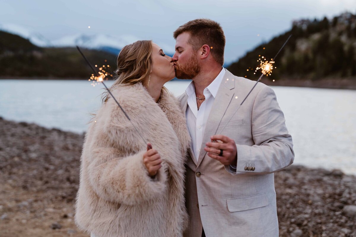 Bride and groom share a kiss while holding sparklers by a mountain lake during their Colorado elopement.