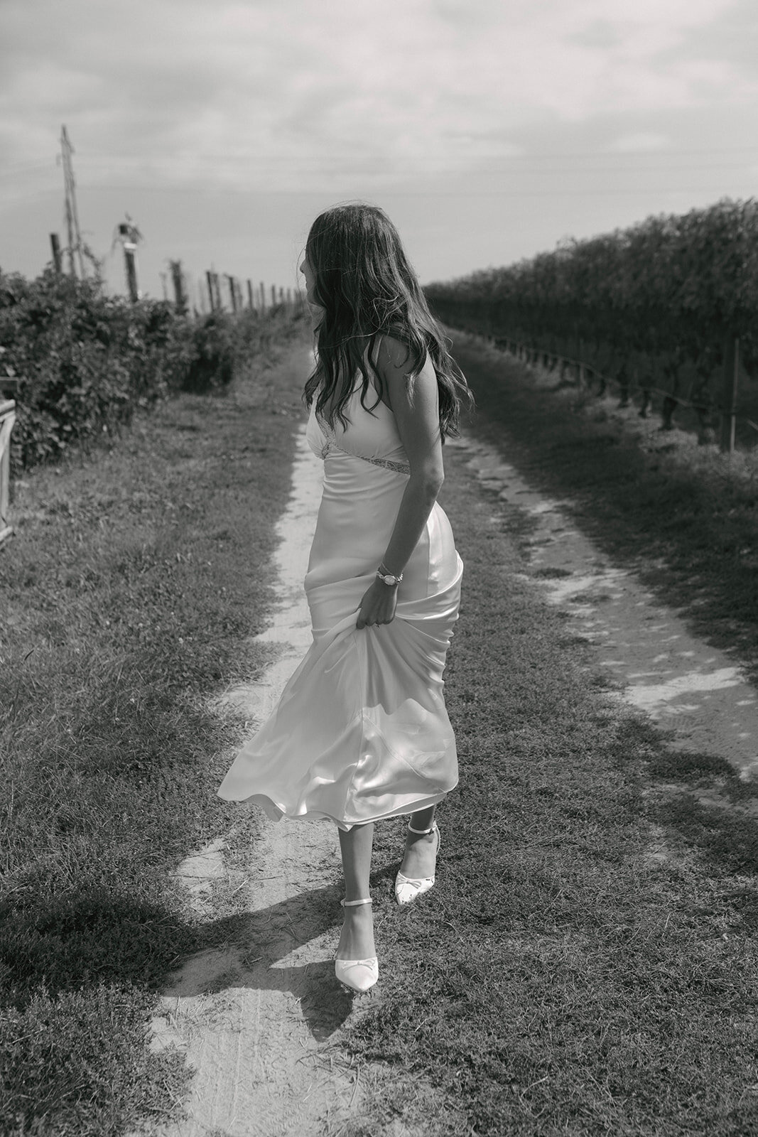 Romantic black-and-white engagement portrait at Rose Hill Vineyards & Inn on the North Fork of Long Island, featuring a woman in a silk dress walking through vineyard rows in an editorial documentary style.
