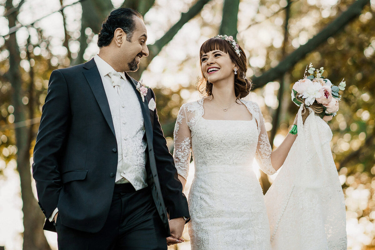 Bride and groom walking hand in hand at golden hour, smiling at each other with soft sunlight glowing behind them.