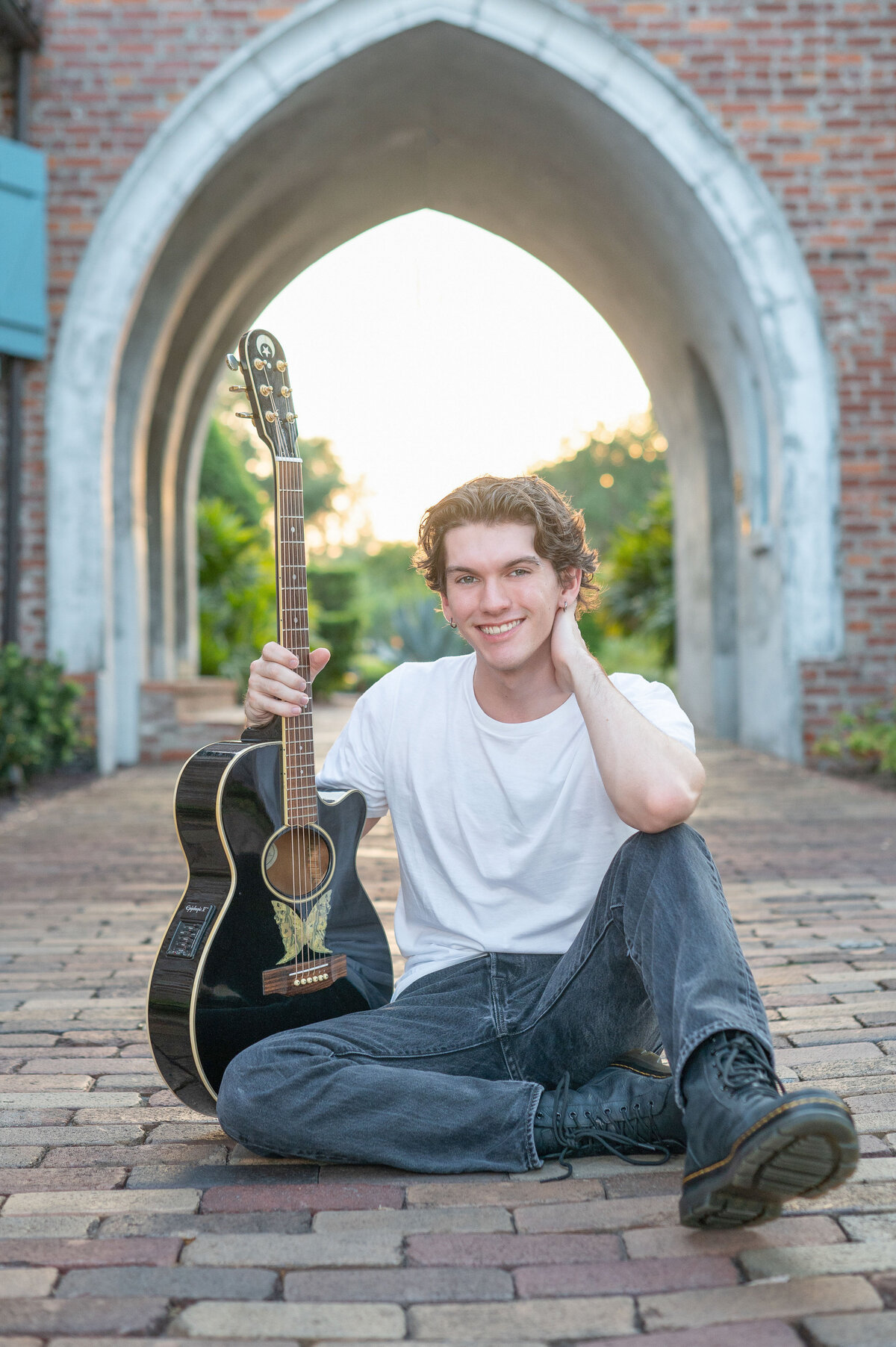 High school senior boy sitting on brick walkway holding a guitar smiling.