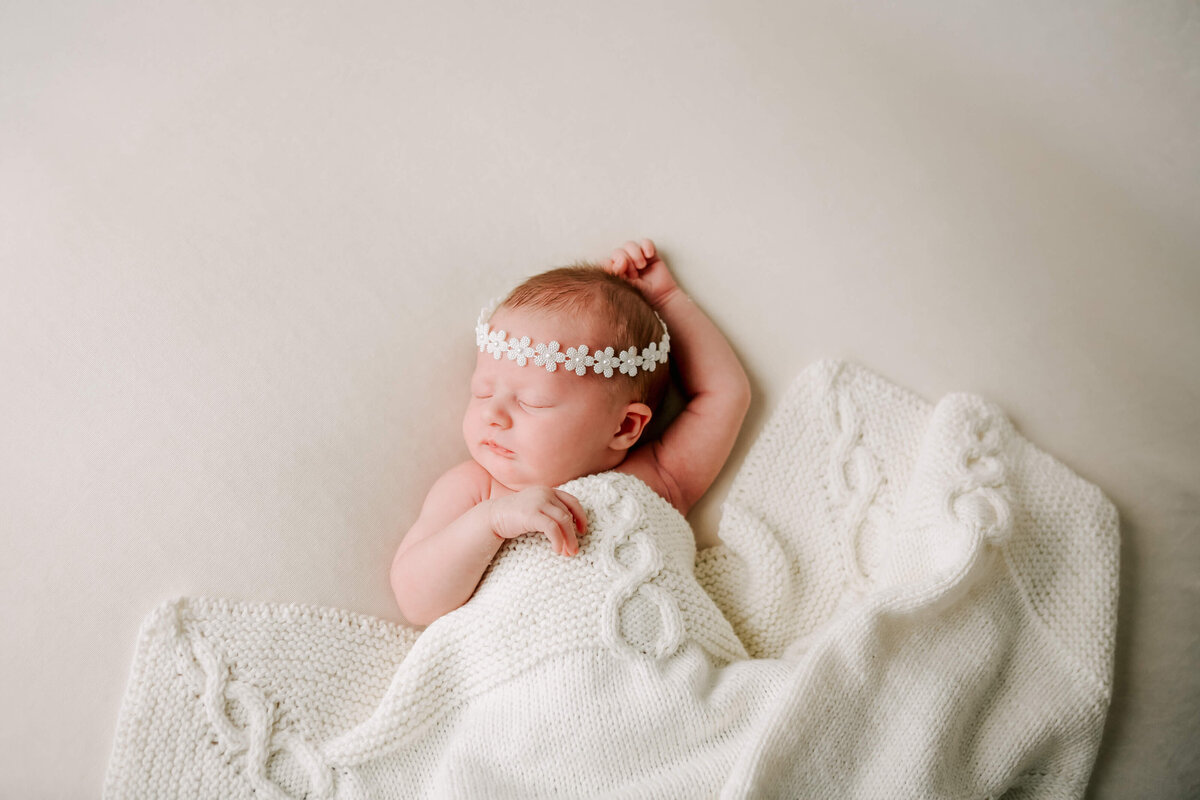 baby girl in white knitted blanket and white beaded flower headband resting on her back with her arm up  in Guelph