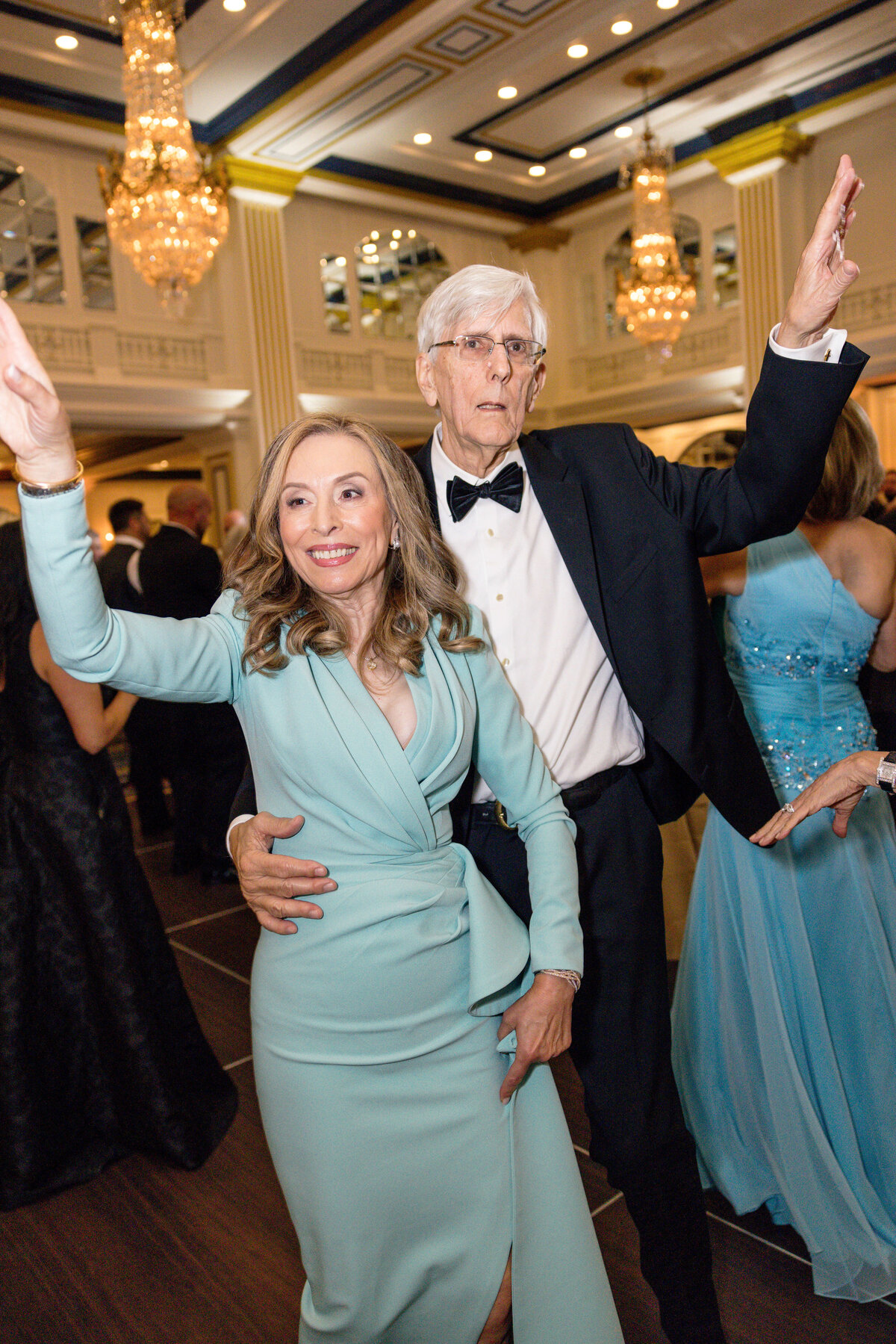 A woman in a light blue dress and a man in a black suit with a bow tie are dancing joyfully at an elegant event. The room is adorned with chandeliers, and other guests in formal attire are in the background.