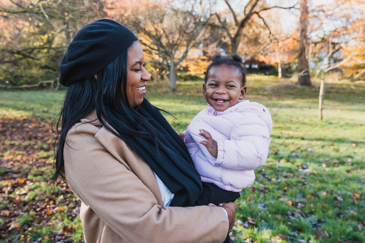 A smiling woman holding a joyful toddler in a park during autumn. both are dressed warmly and the background shows colorful fall leaves.