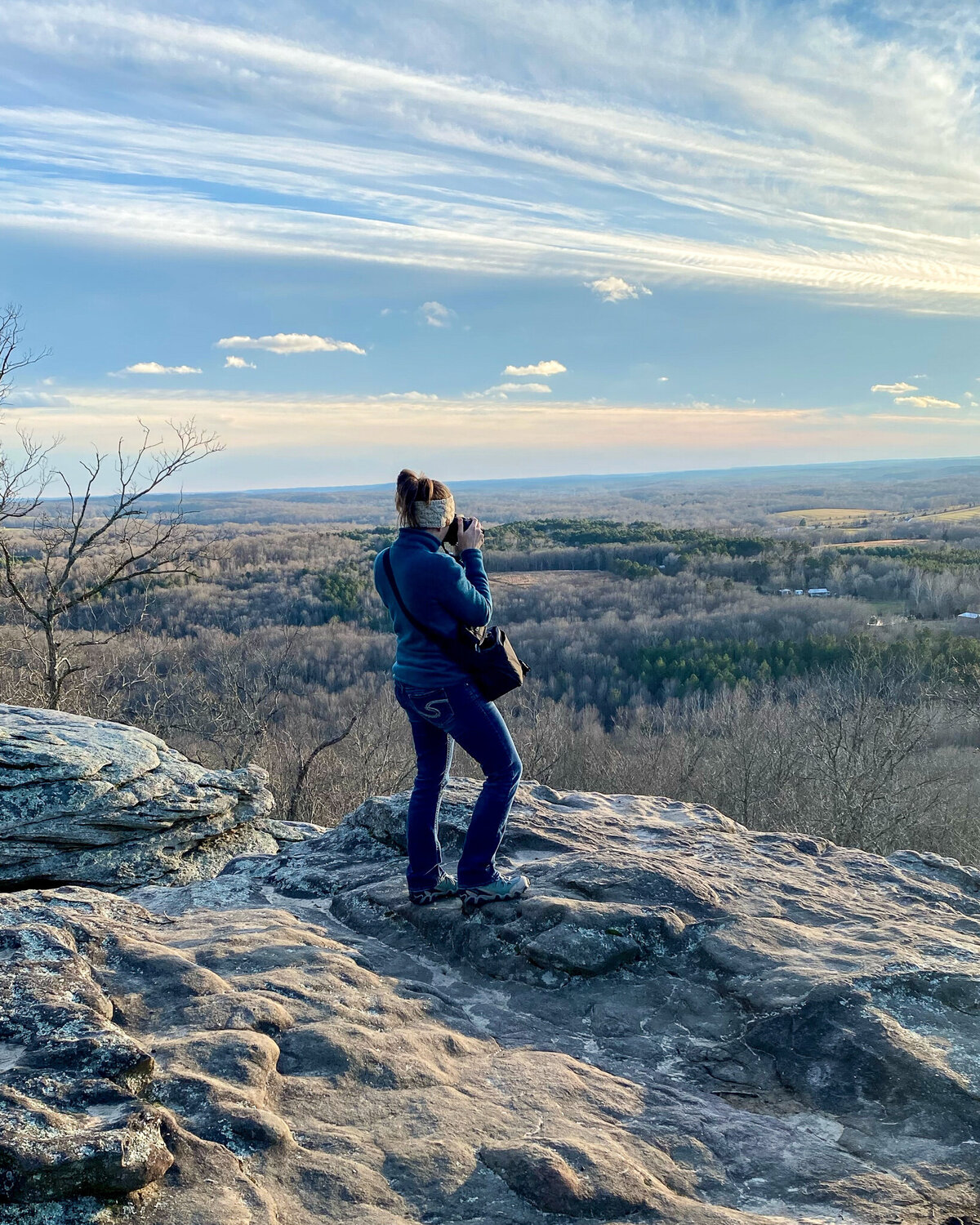 woman standing on a rock formation looking out over a wide area, taking photos during the sunset