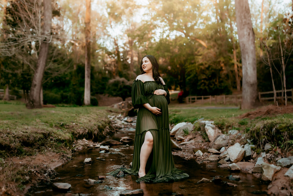 Pregnant woman in a green dress stands in a shallow stream, surrounded by a wooded landscape with tall trees and sunlight filtering through.