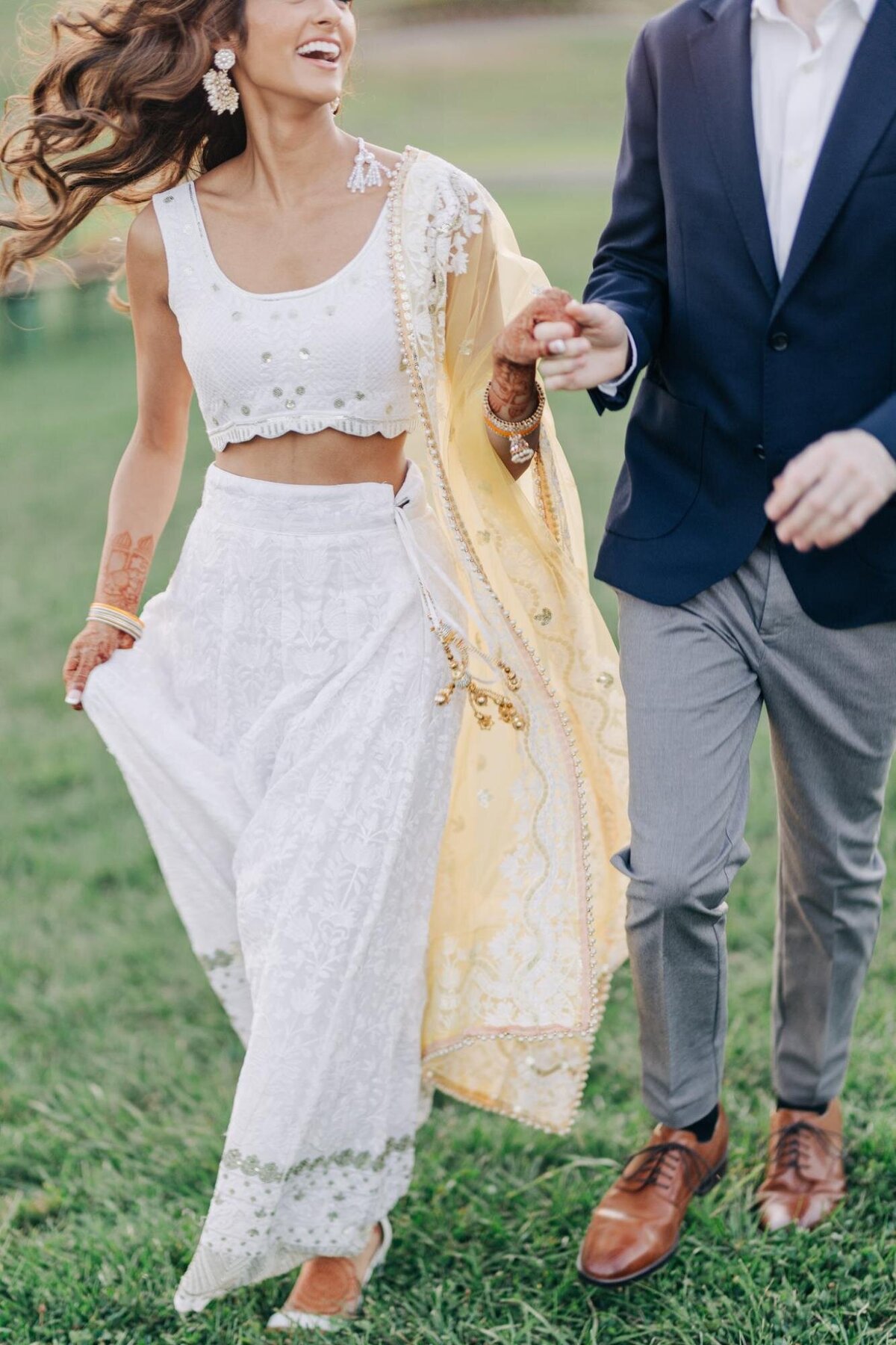 A joyful woman in a white and gold traditional outfit holding hands with a man in a suit, both walking on grass.