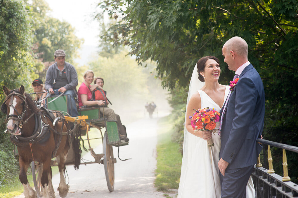 brunette bride wearing a v-neck, ballgown style dress, holding an oramge and pink flower bouquet with her groom wearing a navy suit while standing on a bridge in Killarney National Park with a horse and cart full of people in the background