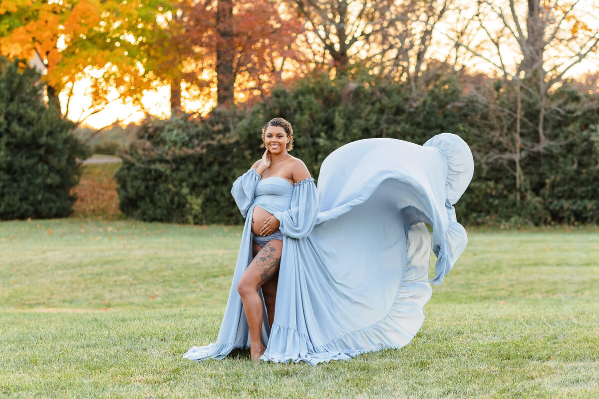 A pregnant woman posing for a photo in Chantilly, VA with her blue dress flying in the wind behind her.