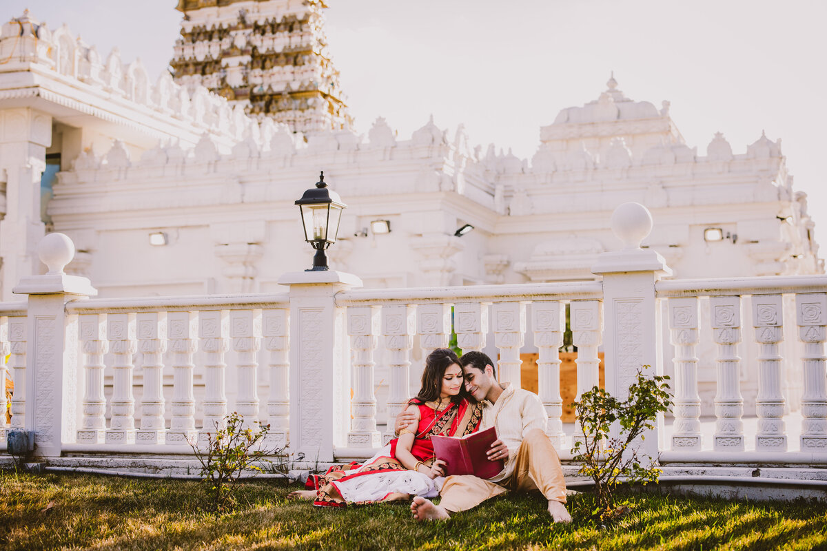 A couple in traditional bengali indian clothing sitting on the grass reading a book during their engagement session at the Sri venkateswara balaji temple in bridgewater nj