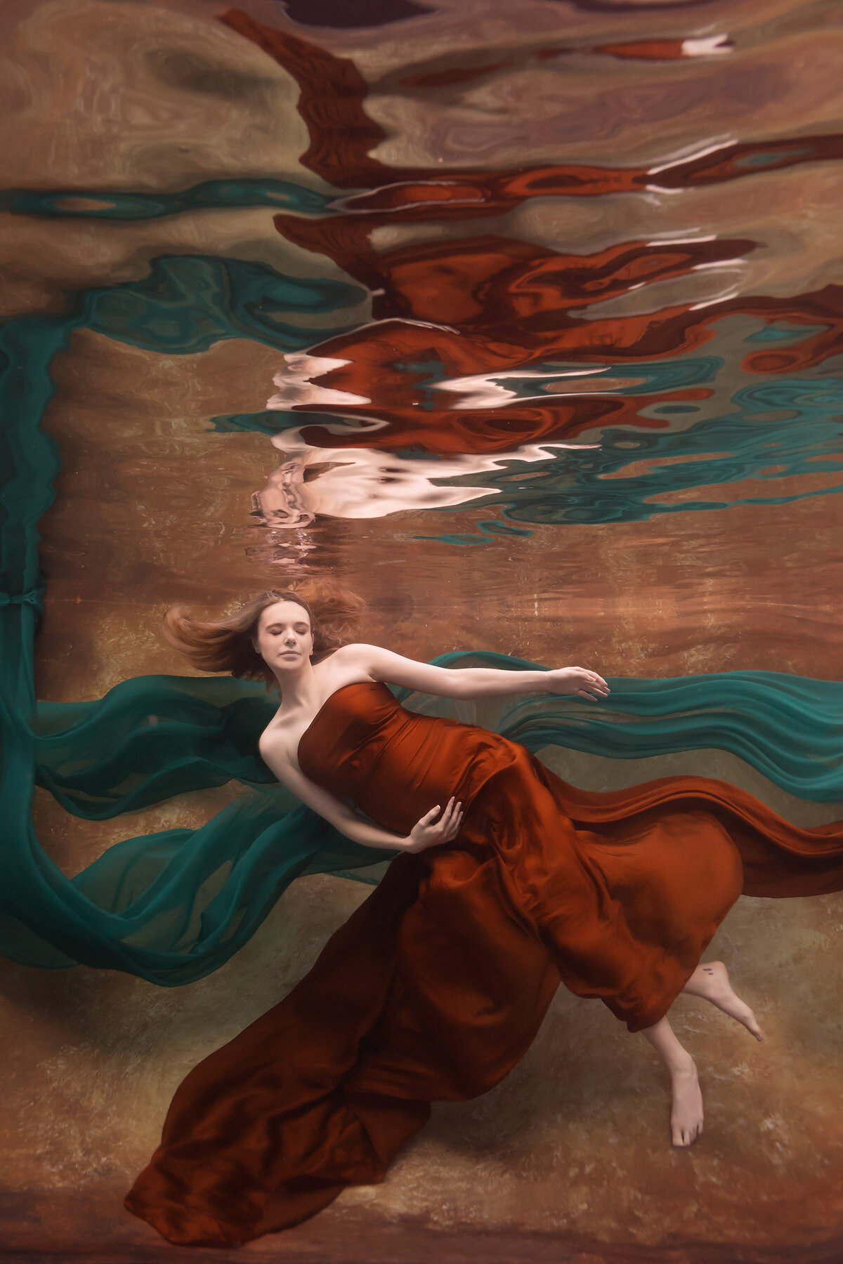 A serene underwater portrait by Lindsay Walden Photography showcasing a woman in a deep rust gown, surrounded by flowing emerald fabric. The luxurious, earthy tones of the water and fabric enhance the ethereal feel of the image, highlighting the elegance of underwater fine art maternity photography.