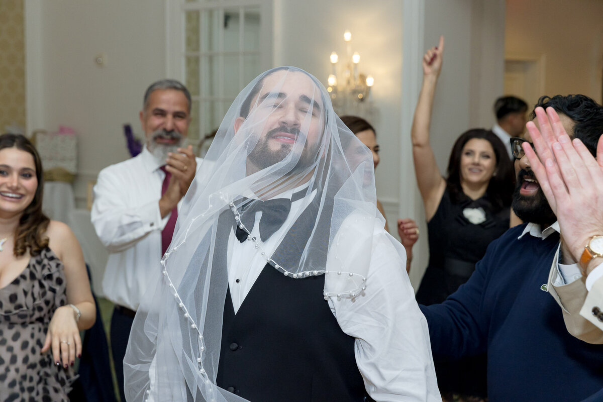 A groom poses for a photo with a veil over his head. Behind him are members of the wedding party clapping and cheering.