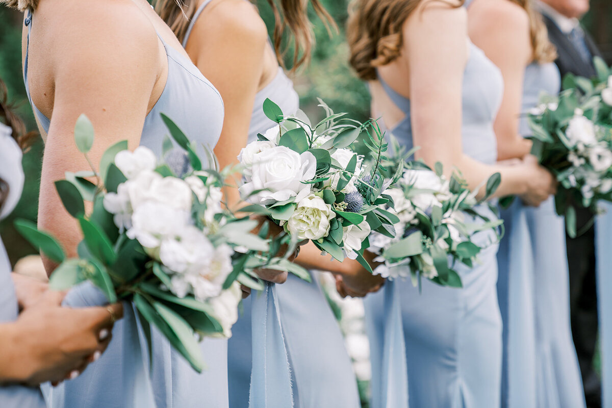 Bridesmaids stand in the wedding ceremony wearing light blue dresses and holding bouquets with white flowers and greenery.