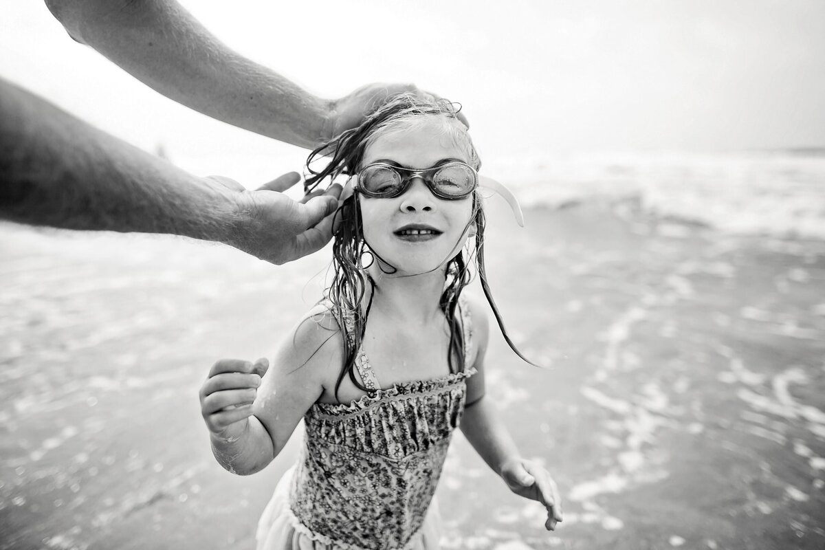 A black and white image of a young girl in her swimsuit getting her  swim googles adjusted.