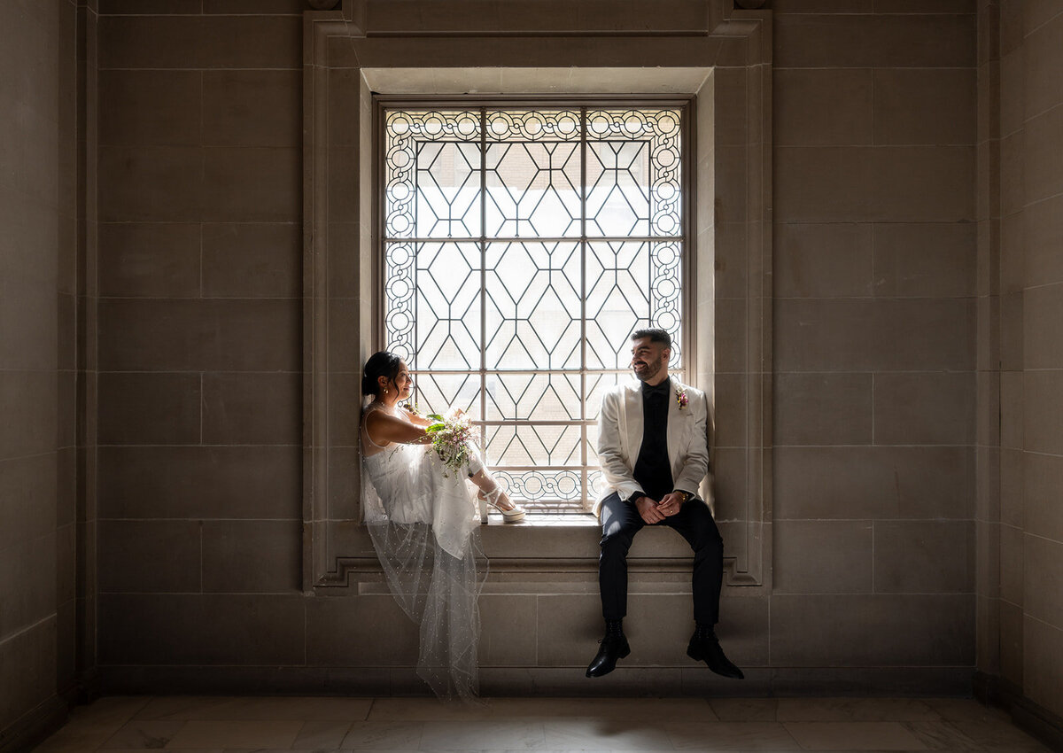 Bride and groom sit inside a large ornamental window sill for a photo at San Francisco City Hall after their wedding ceremony