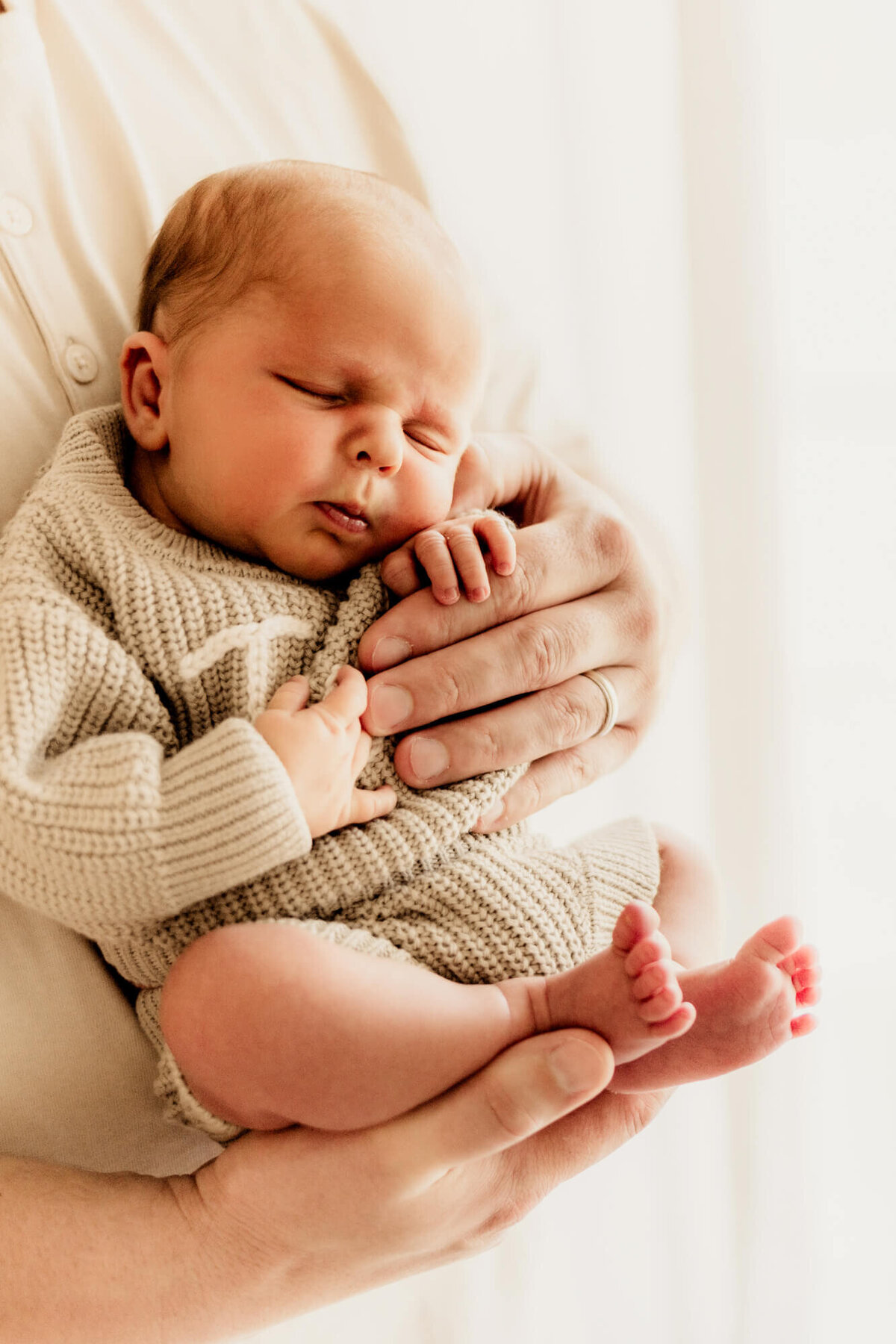 Baby boy in his father's hands with a sage sweater sleeping near a window.