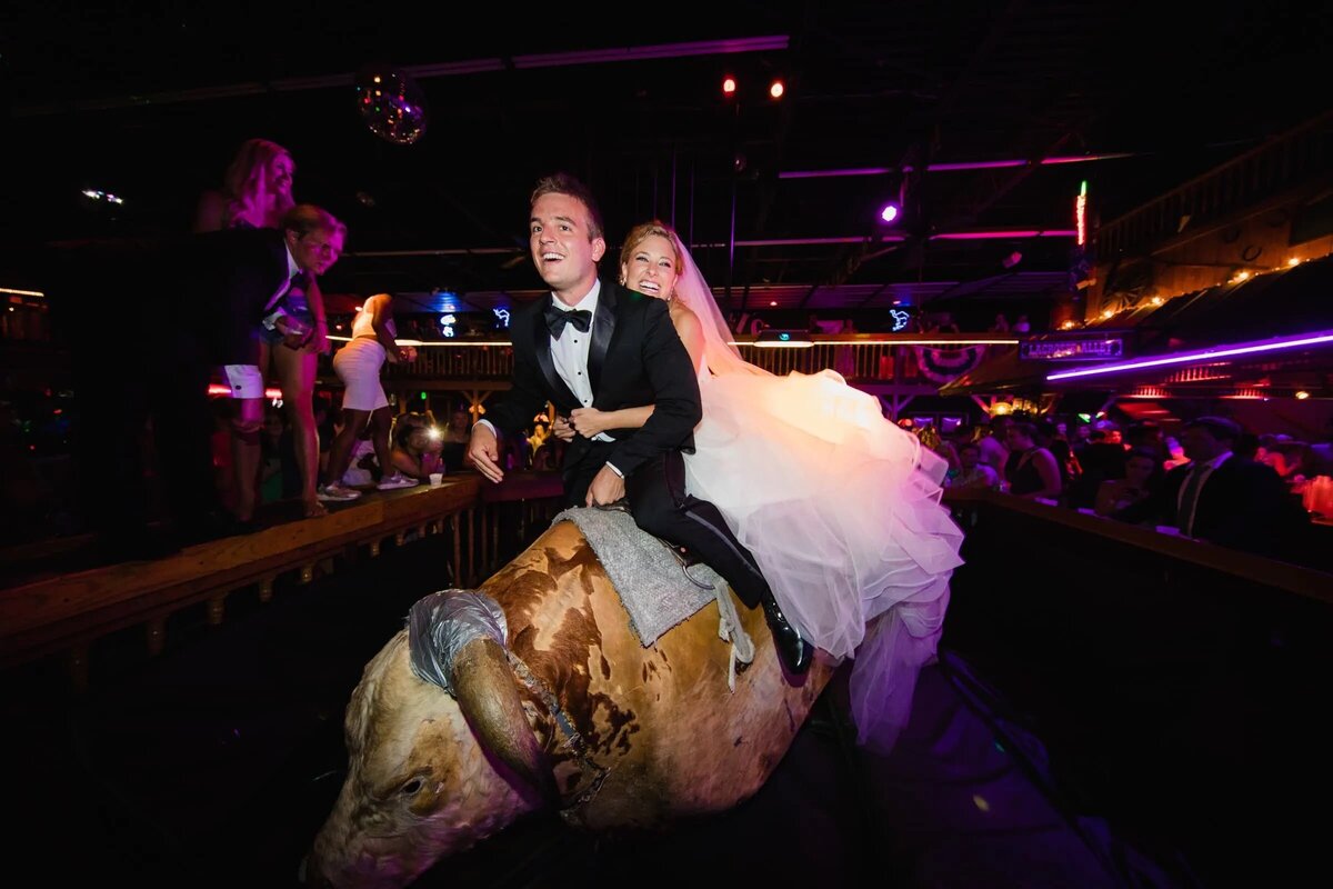 Joyful bride and groom riding a mechanical bull at their wedding reception, with vibrant lighting and guests cheering