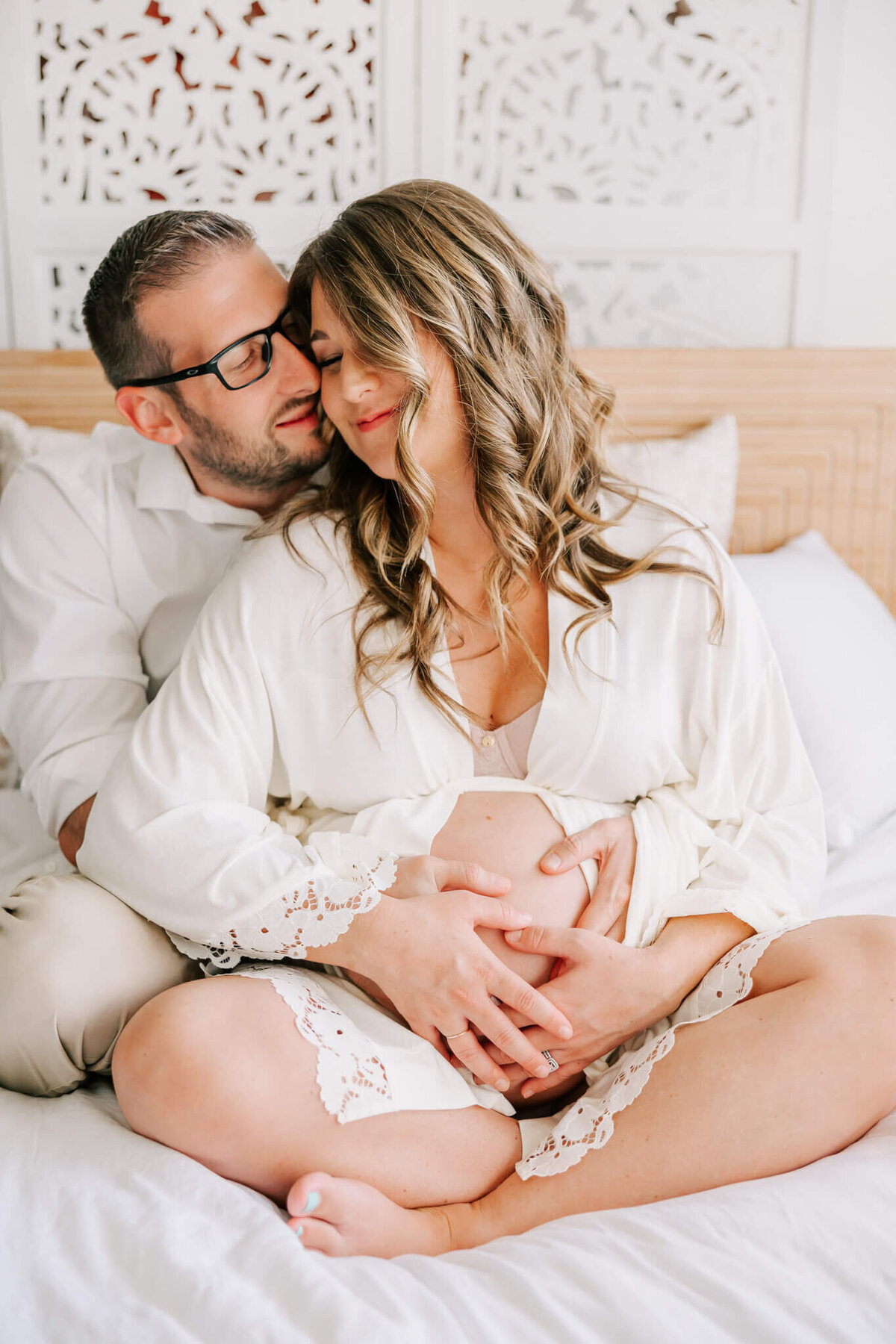 husband and wife sitting on bed with their hands on her belly which is pregnant. She is wearing a white robe.