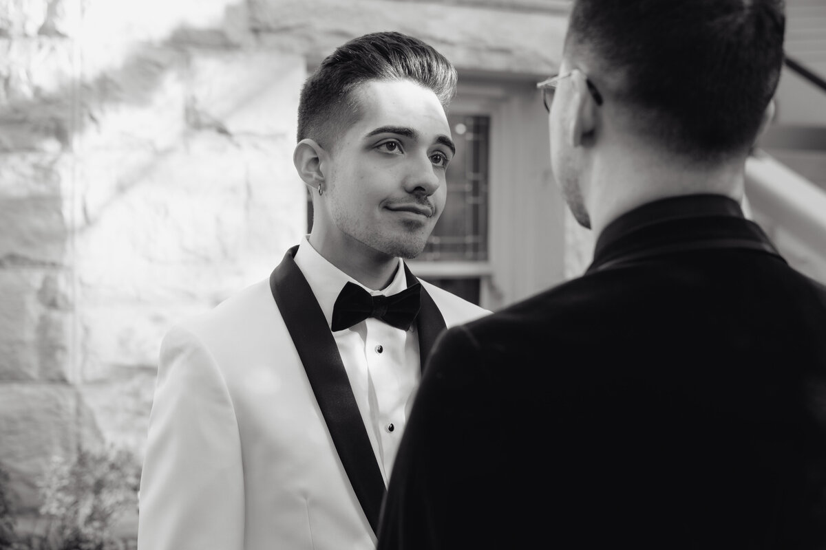 A groom gazes at his partner with emotion, dressed in a black and white tux, captured in a high-end documentary wedding portrait.