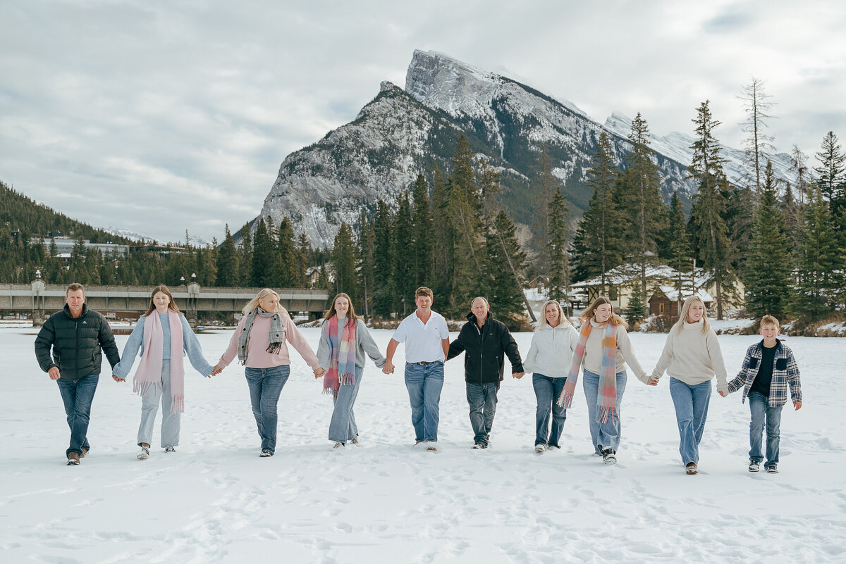 Extended family walks hand in hand across snowy field at Bow River in Banff.