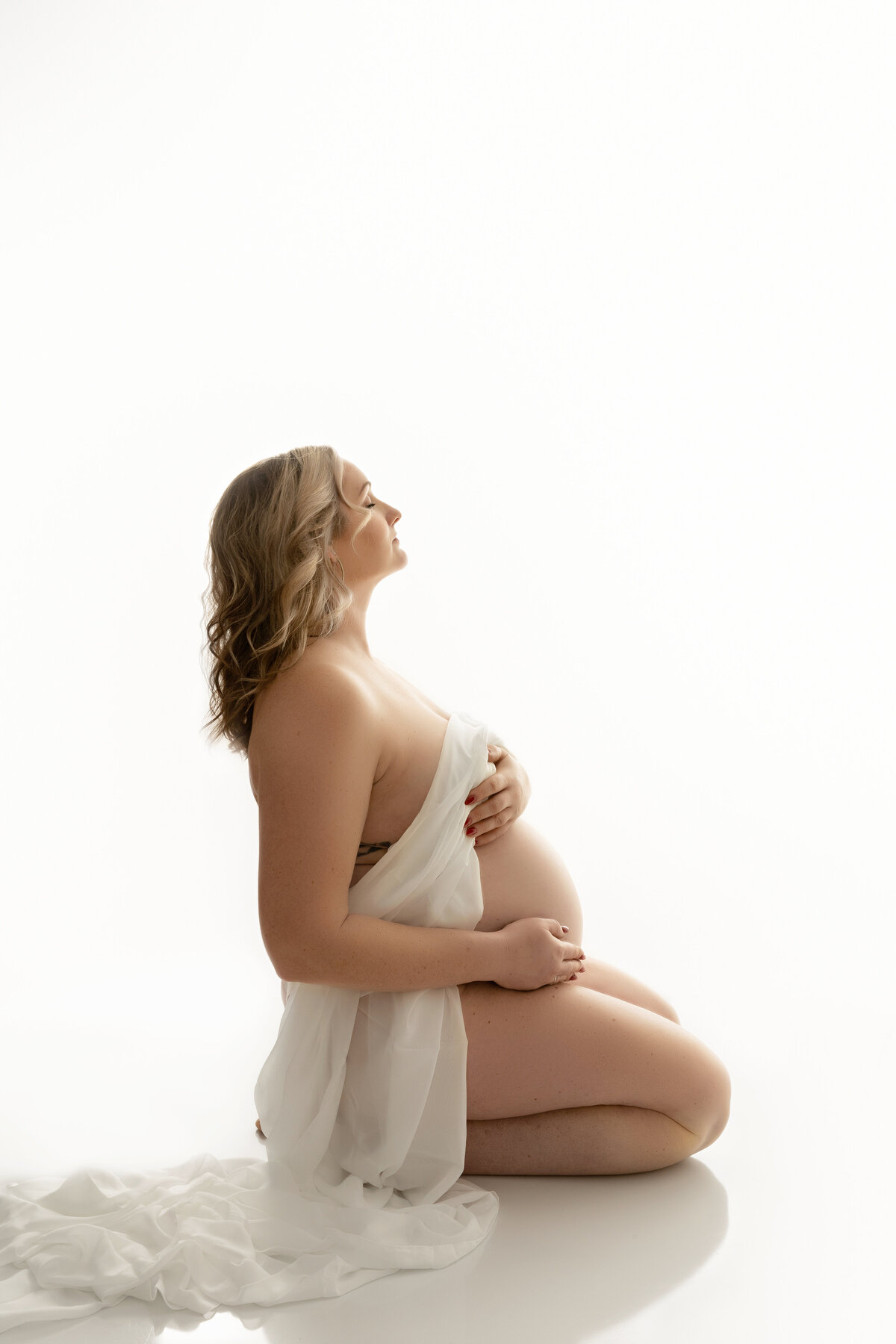 A pregnant woman kneels on the floor of a white studio while covering herself with a long white sheet