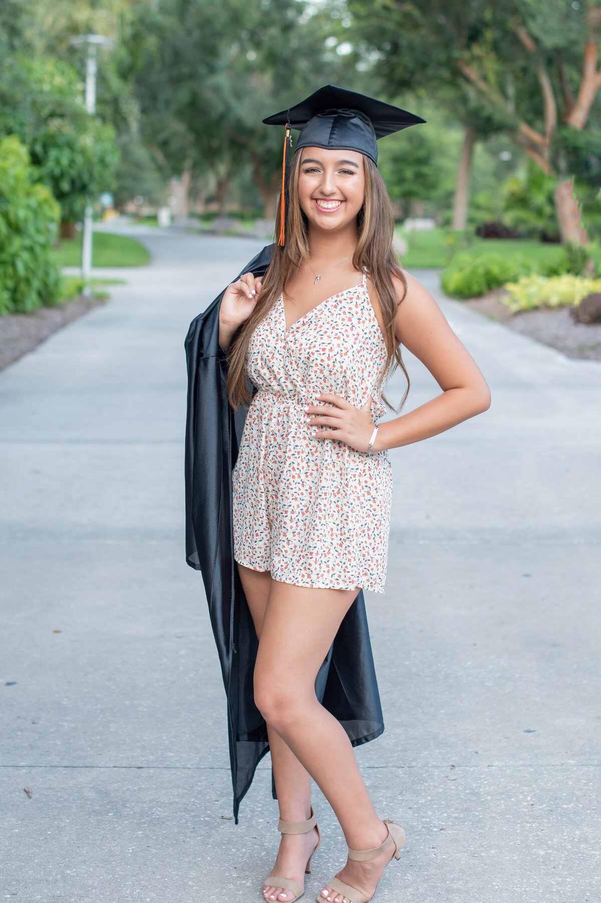 High school senior girl wearing cap holds gown over shoulder while smiling at camera.