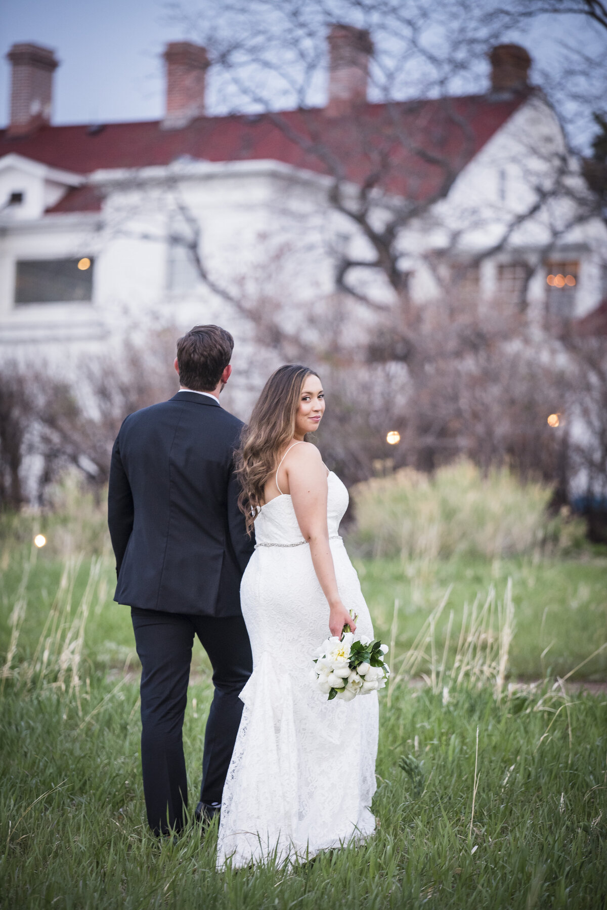 A bride and groom walk away from the camera and the bride glances over her shoulder and gives the camera a soft smile.