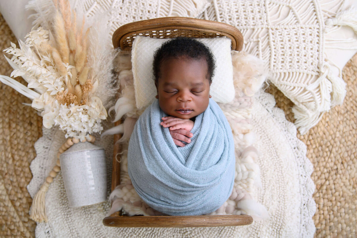 Black newborn boy wrapped in blue laying in a tiny bed for his Burlington newborn photography session.