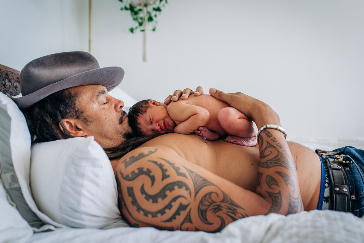 dad with tattoos wearing a hat and laying on bed with newborn in fetal position sleeping on his chest