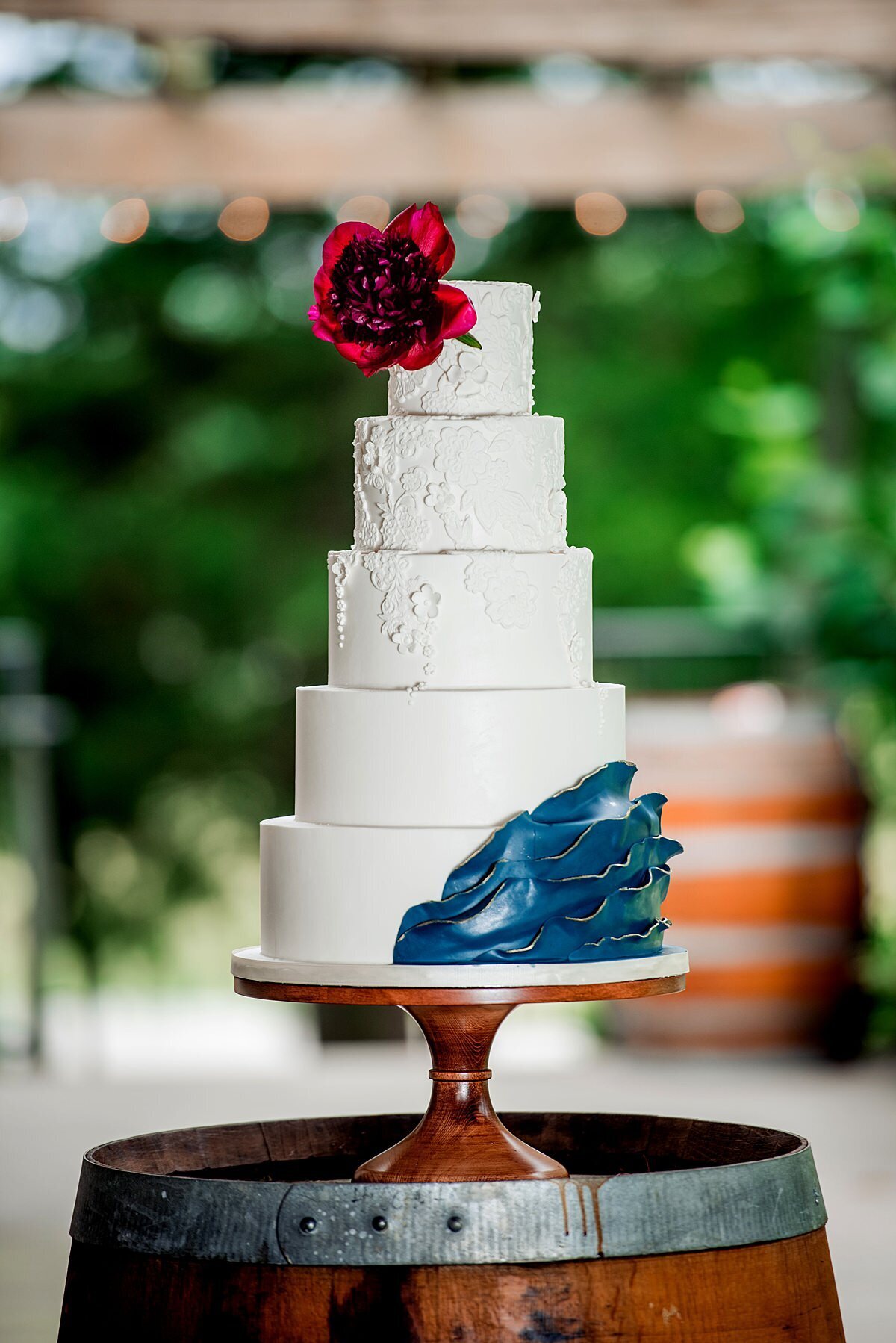A five tier white wedding cake with hand piped lace texture to match the lace on the bodice of the wedding gown accented by teal ruffles made from hand molded chocolate and a large burgundy peony topper. The cake sits on a wooden footed cake stand on top of a wine barrel at Arrington Vineyards.