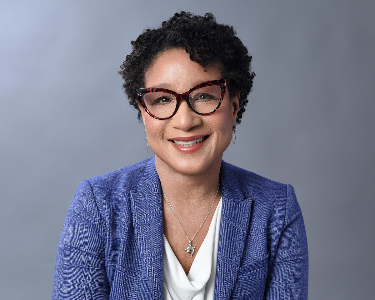 Professional headshot of an individual wearing a stylish blue blazer over a white blouse, complemented by a delicate necklace. The subject’s poised expression against a clean light gray backdrop conveys confidence and warmth—perfect for business branding and professional photography
