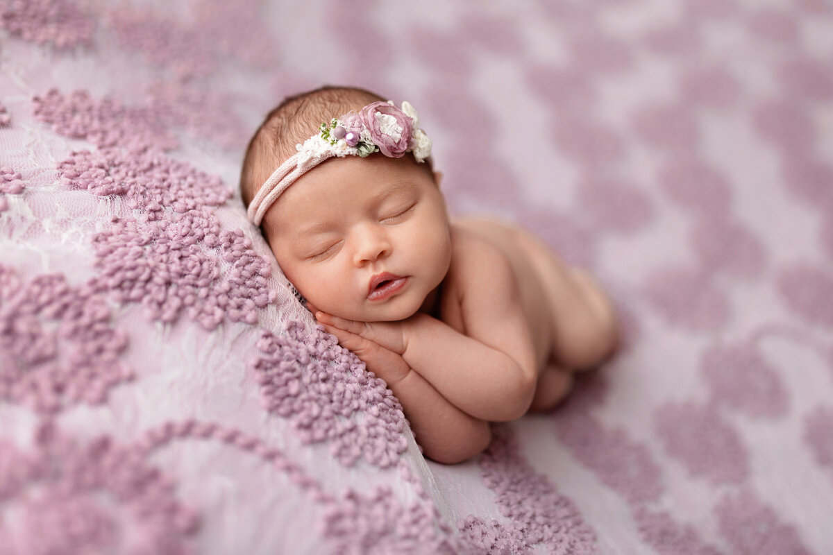 Newborn girl lying on her side on a soft purple patterned blanket wearing a floral headband.