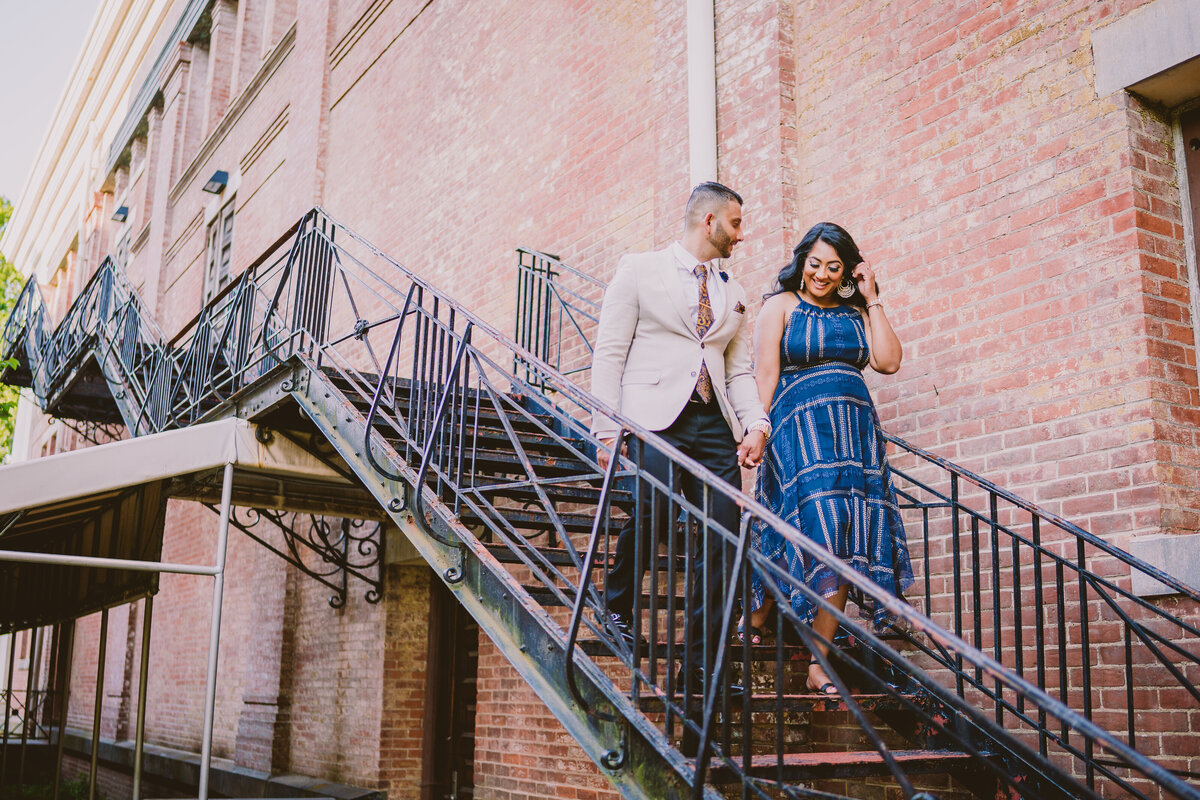 A couple during their engagement shoot walking down metal stairs attached to the outside of a building at snug harbor staten island ny