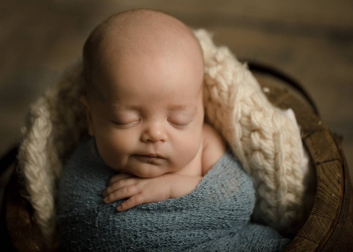 Newborn boy in a light blue wrap sitting inside of a wooden bucket.
