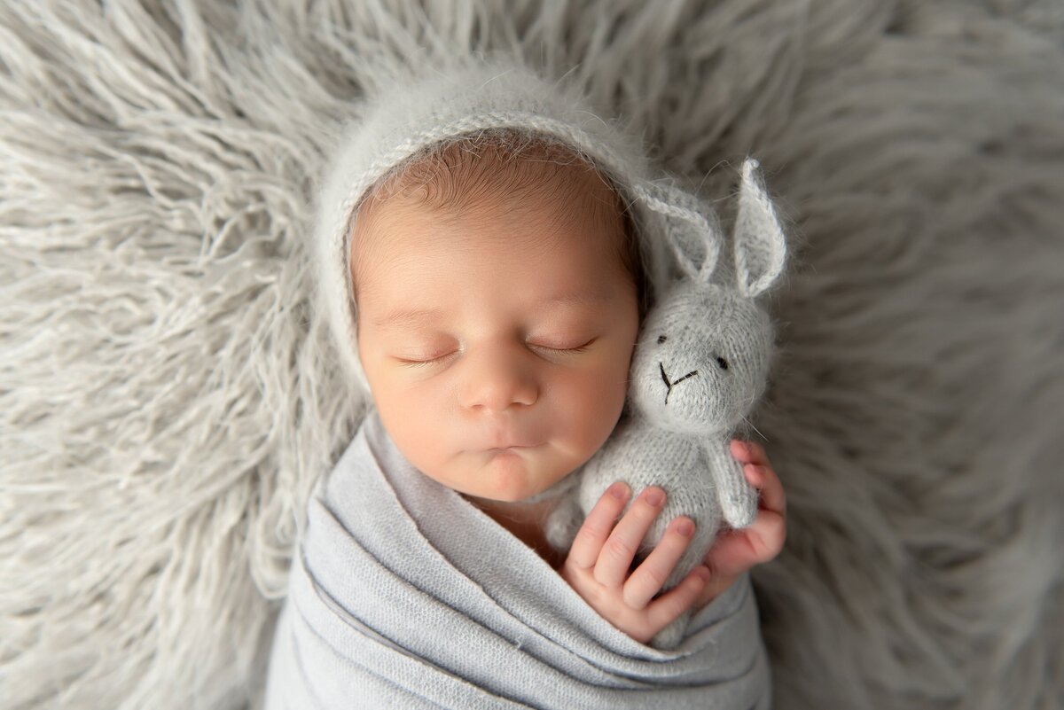 Newborn pictures near me will showcase baby boy in gray wrap and bonnet holding a bunny.