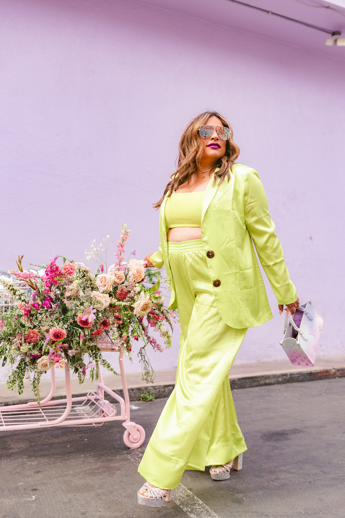 Creative branding portrait of a confident woman in a chartreuse suit and platform heels posing with a flower-filled shopping cart in front of a pastel purple wall — vibrant and fashion-forward.
