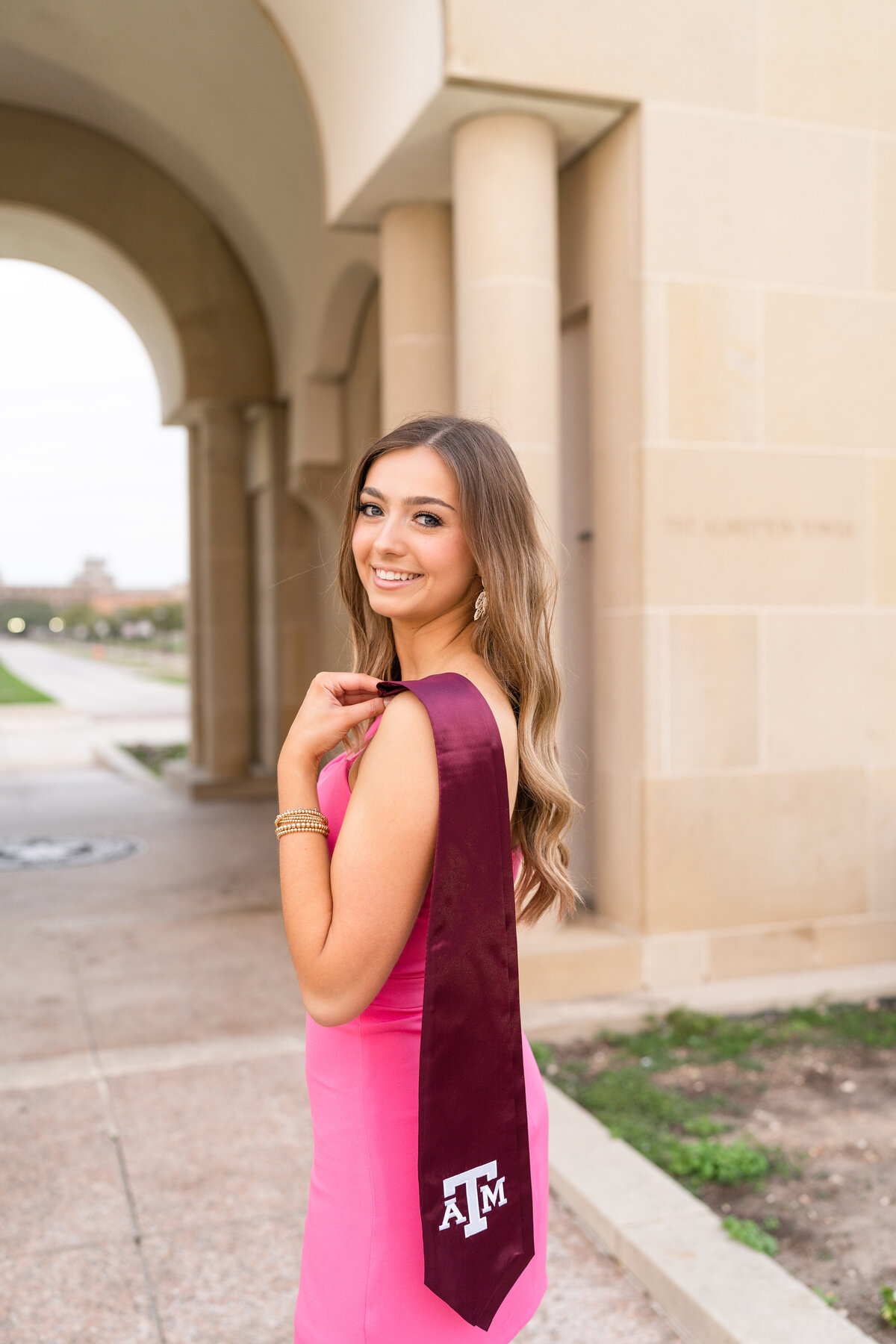 Texas A&M senior girl smiling over shoulder while holding maroon stole and wearing pink dress at the Bell Tower