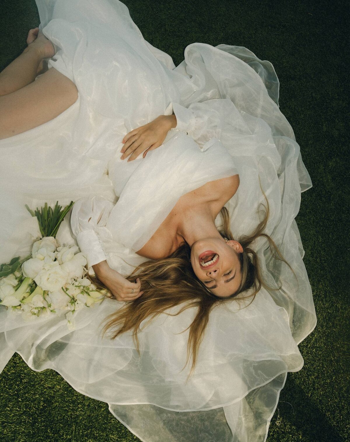 Bride laughing while lying on grass in a flowing tulle gown with white bouquet at a Houston wedding.