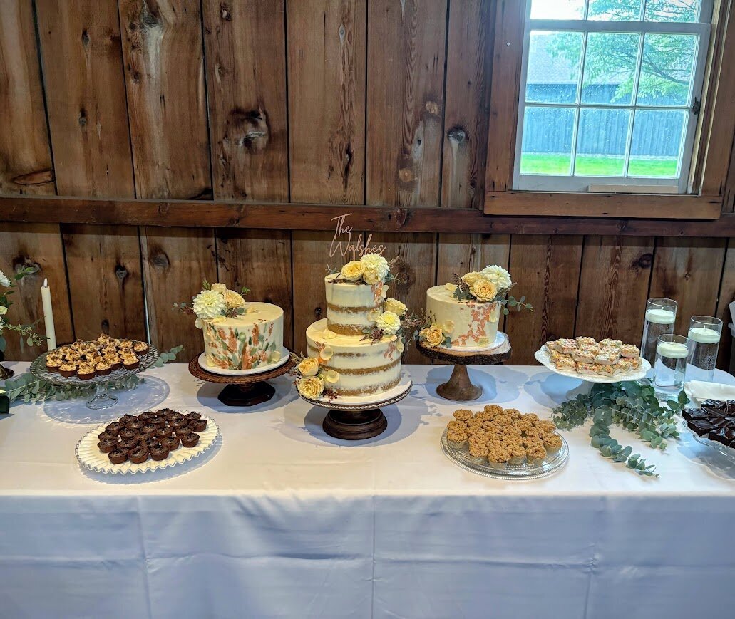A table holds a three tier wedding cake display, surrounded by other stands of mini pastries. The cakes are all in shades of white, terracotta, and green with matching florals. The mini pastries include peanut butter tarts, fudge brownies, apple pies, and rice krispie treats.