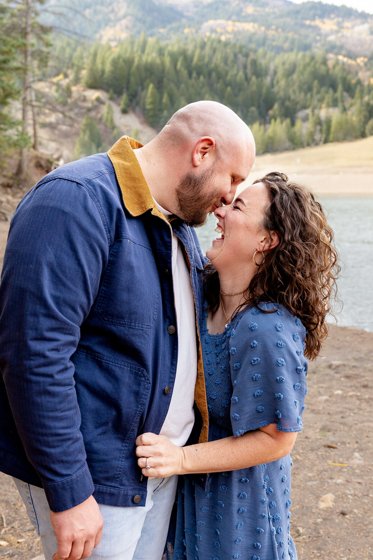 A couple stands close, smiling and touching noses, sharing a joyful moment with a stunning mountain and lake backdrop at Tibble Fork Reservoir.