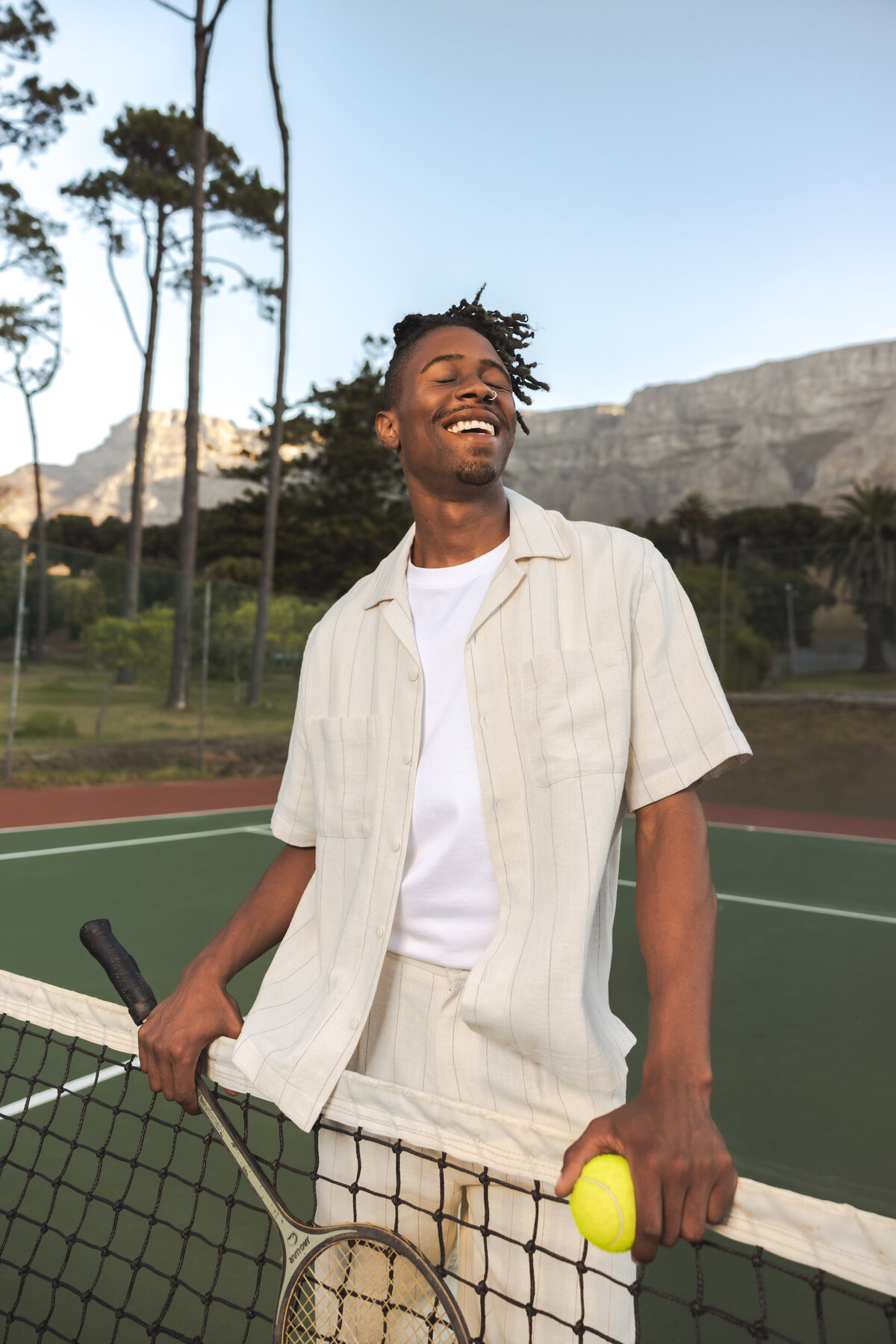 Man smiling with eyes closed while leaning on a tennis net, holding a racket and tennis ball, with Table Mountain in the background.