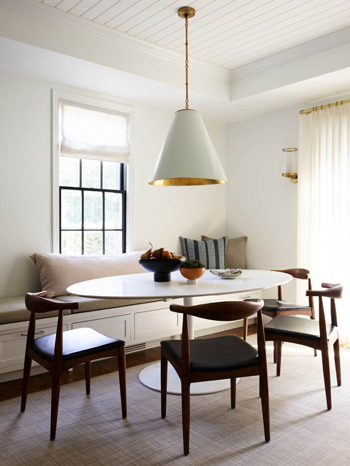 Explore the casual dining room in a historic Hingham home, with a custom bench under the window, Saarinen-inspired dining table, mid-century furniture, and white linen drapery.