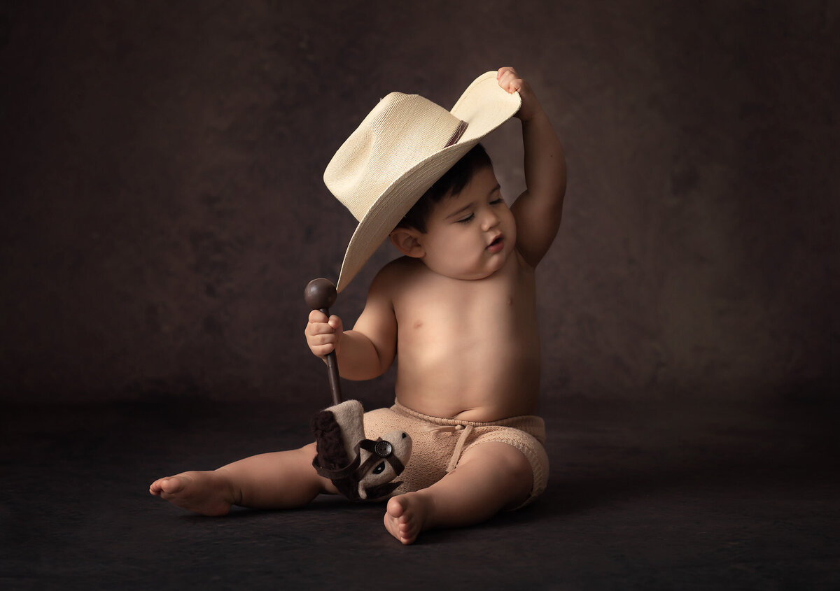 Adorable baby wearing a cowboy hat, holding a small plush horse toy while sitting on a dark, rustic background. This charming milestone portrait captures the playful innocence of childhood, perfectly created by a talented Dallas baby milestone photographer.