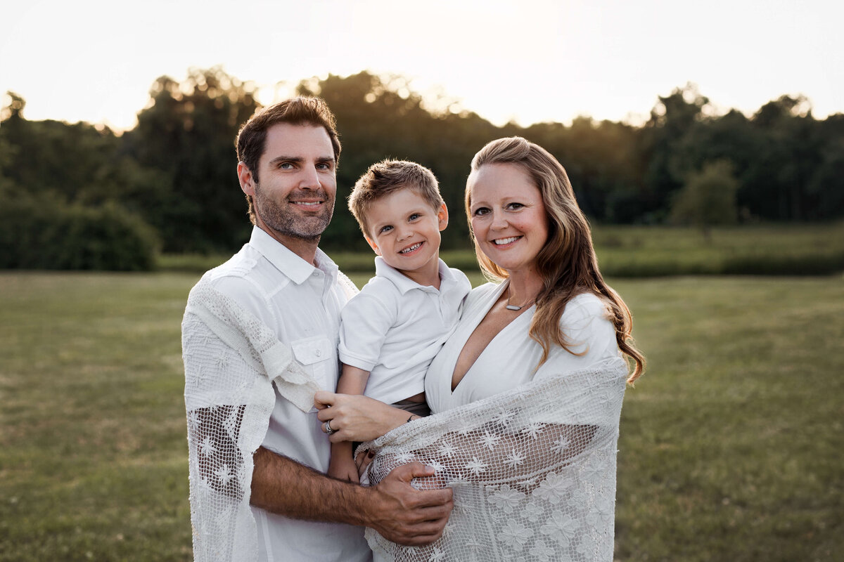 mom and dad smiling with their 6 year old son in the middle , wrapped in a blanket at sunset in the summer
