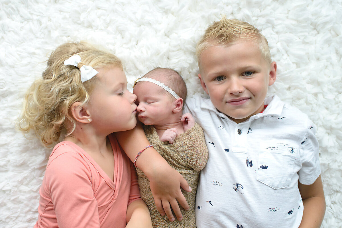 Siblings holding their new baby sister.  Older sister is kissing baby on the nose while older brother smiles at the camera.