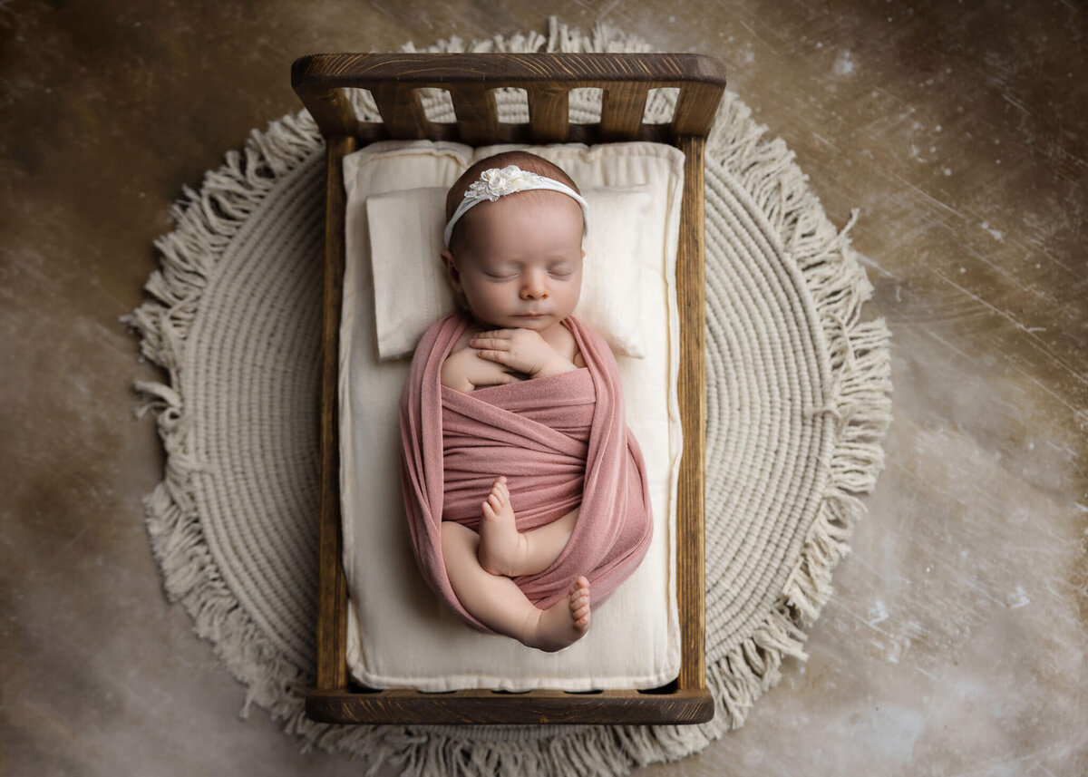 newborn baby wrapped in pink fabric wearing a white floral headband asleep on a tiny wooden bed