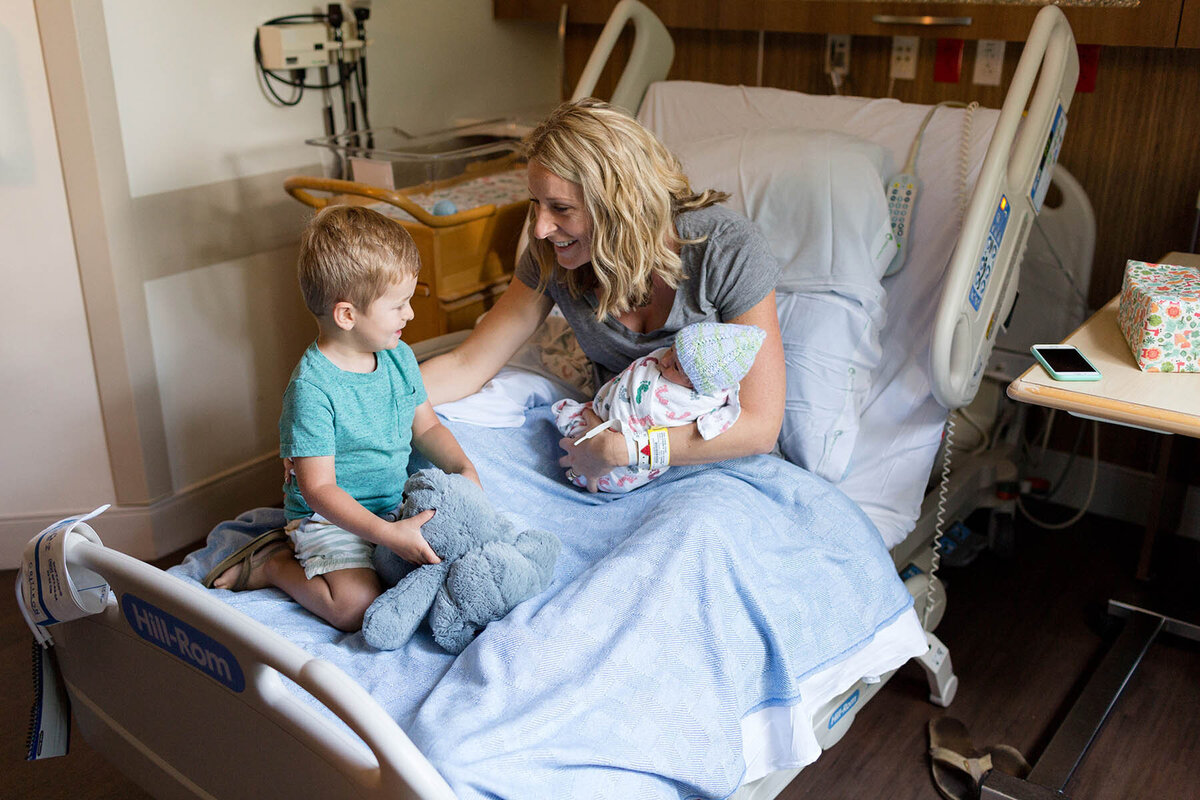 a mother sits on a hospital bed holding her newborn baby and smiling at her toddler son